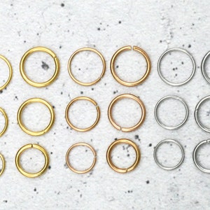 Puede incluir: Un conjunto de 18 aros para pendientes de oro, oro rosa y plata. Los aros están dispuestos en tres filas de seis, con cada fila presentando un color de metal diferente. Los aros son de diferentes tamaños, con los aros más pequeños en la fila inferior y los aros más grandes en la fila superior.