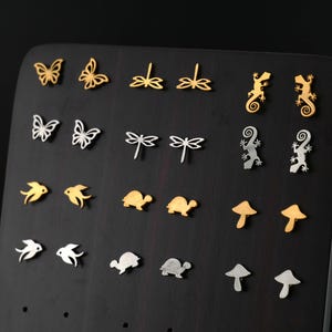 May include: A display of silver and gold earrings with various designs, including butterflies, dragonflies, turtles, mushrooms, and lizards.