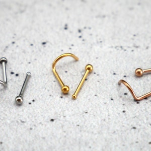 May include: Three nose rings with different styles and colors. One is a silver hoop, one is a gold straight bar, and one is a rose gold curved bar.