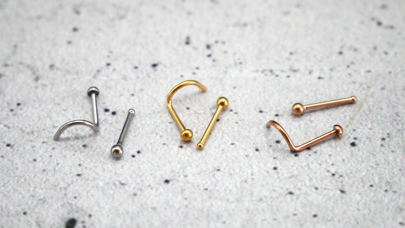 May include: Three nose rings with different styles and colors. One is a silver hoop, one is a gold straight bar, and one is a rose gold curved bar.