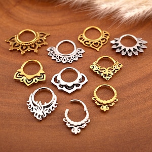 Pode incluir: Uma coleção de piercings de nariz ornamentados em tons de ouro e prata. Os designs incluem padrões florais, geométricos e com contas. As joias são exibidas em uma superfície marrom, mostrando os detalhes intrincados de cada peça.
