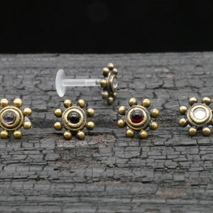 Op de afbeelding: Vijf gouden neusringen met verschillende gekleurde edelstenen in het midden. De ringen hebben de vorm van een zon met een cirkel in het midden. De edelstenen zijn zwart, rood, helder en iriserend.