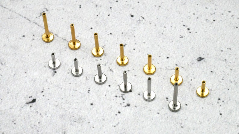May include: A set of 10 gold and silver body jewellery pieces. The jewellery is in the shape of a small stud with a flat base and a short, straight post. The jewellery is arranged in two rows on a white surface.