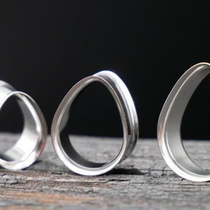 May include: Three silver teardrop-shaped ear gauges of different sizes. The gauges are made of metal and have a smooth, polished finish.