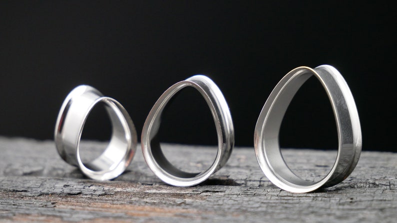 May include: Three silver teardrop-shaped ear gauges of different sizes. The gauges are made of metal and have a smooth, polished finish.