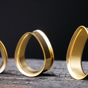May include: Three gold teardrop-shaped ear gauges of different sizes. The gauges are made of metal and have a smooth, polished finish.