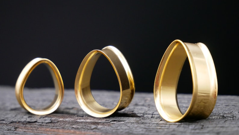 May include: Three gold teardrop-shaped ear gauges of different sizes. The gauges are made of metal and have a smooth, polished finish.