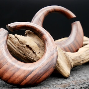 May include: Two wooden hoop earrings with a dark brown and reddish brown wood grain pattern. The earrings are displayed on a piece of driftwood.