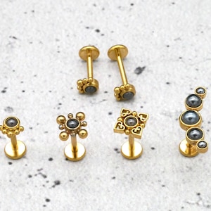 Könnte beinhalten: Sechs goldene Körperschmuckstücke mit schwarzen Edelstein-Akzenten. Der Schmuck verfügt über verschiedene Designs, darunter einen einfachen runden Stein, einen quadratischen Stein mit dekorativem Rand und eine geschwungene Reihe von Steinen.