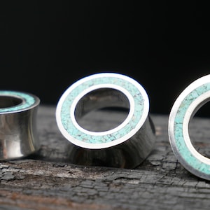 May include: Three silver ear gauges with turquoise inlay. The gauges are different sizes and are arranged in a row on a dark wooden surface.