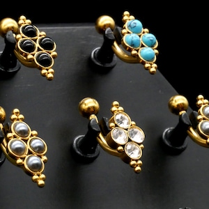 Puede incluir: Cinco piezas de joyería corporal de oro con piedras preciosas de diferentes colores. Cada pieza tiene una barra curva con una bola en un extremo y un grupo de piedras preciosas en forma de diamante en el otro extremo. Las piedras preciosas son negras, turquesa, transparentes y grises.