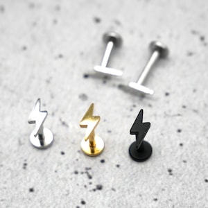 Puede incluir: Tres pendientes de botón con forma de rayo en plata, oro y negro. Los pendientes están sobre un fondo gris.