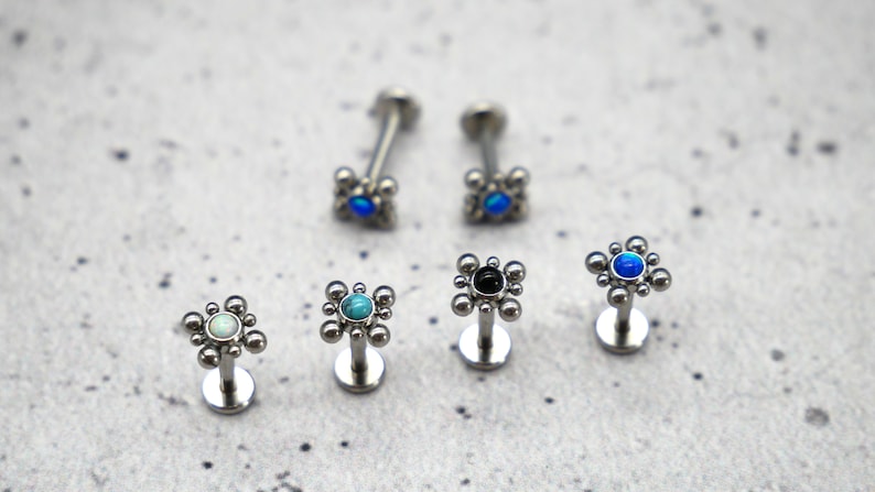 May include: Six different styles of surgical steel body jewelry with various colored gemstones. The jewelry is in the shape of a small square with a round gemstone in the center.