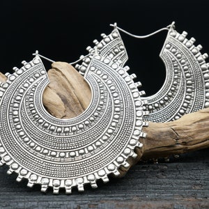 Puede incluir: Un par de grandes pendientes de aro plateados con intrincados diseños circulares. Los pendientes están hechos de metal y tienen un acabado brillante.