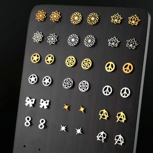 Op de afbeelding: Een display met verschillende zilveren en gouden oorbellen, waaronder sterren, vredestekens, bloemen en anarchie symbolen. De oorbellen zijn gerangschikt op een zwart displaybord.