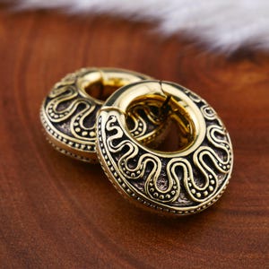 Può includere: Due orecchini circolari color oro con intricati motivi a vortice marrone scuro. Gli orecchini hanno una chiusura a cerniera e sono esposti su una superficie di legno. Il design ricorda i gioielli antichi o tribali.
