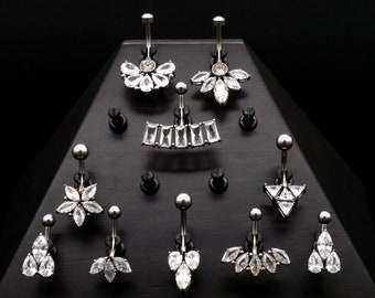 May include: A display of various belly button rings with clear crystal accents. The rings are arranged on a black triangular stand.