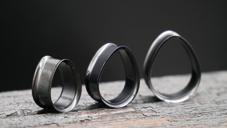 May include: Three black and silver ear gauges of different sizes. The gauges are shaped like ovals with a slightly pointed top and bottom.