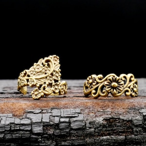 Könnte beinhalten: Zwei goldfarbene Ringe mit filigranen Blumenmustern. Die Ringe liegen auf einer dunklen Holzoberfläche.