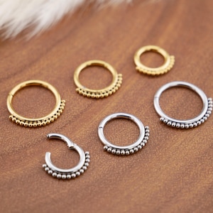 Könnte beinhalten: Eine Auswahl an goldenen und silbernen Septumringen mit einem Perlendesign. Die Ringe sind kreisförmig und haben einen Klappverschluss für einfaches Einsetzen. Der Schmuck wird auf einer braunen Oberfläche präsentiert.