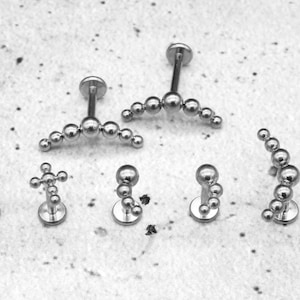 Puede incluir: Seis piezas diferentes de joyería corporal plateada con varios diseños, que incluyen una línea curva de cuentas, una cruz y un grupo de cuentas.