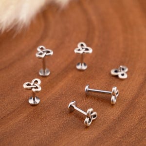 Op de afbeelding: Een collectie labret-piercings van zilverkleurig metaal met een triquetra-knoopontwerp. Elk sieraad bestaat uit een platte ronde basis, een rechte staaf en een decoratieve bovenkant in de vorm van een gevlochten knoop met drie punten.