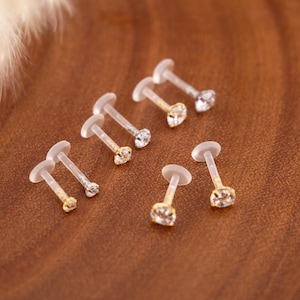 Op de afbeelding: Een verzameling van zes labret-piercings met doorzichtige, flexibele kunststof staafjes en platte, ronde achterkantjes. Elke piercing is voorzien van een kleine, ronde, heldere gefacetteerde edelsteen in een goudkleurige metalen zetting.