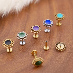 Può includere: Una collezione di piercing labret a base piatta con centri circolari in nero, verde, viola, blu e turchese. Ogni gioiello presenta una cornice metallica decorativa a forma di fiore smerlato attorno alla pietra e un perno filettato.