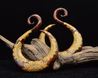 May include: A pair of wooden ear gauges with intricate gold designs. The gauges are shaped like a crescent moon and have a curved hook at the top.