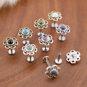 May include: A collection of flower-shaped labret studs featuring ornate metal petals in gold and silver tones. Each stud is centered with a small, smooth, round gemstone in various colors including turquoise, black, green, and iridescent white.