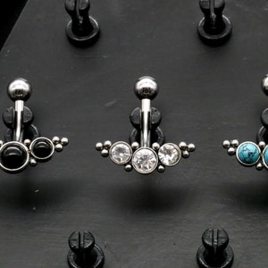 May include: Three belly button rings with different coloured stones. The first ring has three black stones, the second ring has three clear stones, and the third ring has three turquoise stones.