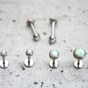 Puede incluir: Cinco piezas de joyería corporal plateadas con piedras que parecen opales. Las piezas están dispuestas en fila sobre una superficie gris texturizada. Las piedras son blancas con un toque de azul y verde.