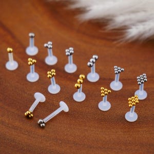 May include: A set of 14 clear plastic body jewelry pieces with silver and gold ball accents. The jewelry pieces are arranged on a wooden surface.
