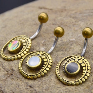 Puede incluir: Tres piercings para el ombligo con un diseño en espiral de color dorado y una barra curva plateada. Cada anillo presenta una piedra central diferente: iridiscente, blanca y negra. La bola superior es dorada.