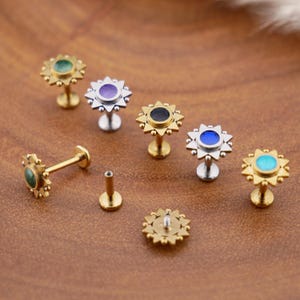 May include: A collection of flat-back stud earrings featuring a sunburst design with small spheres around the perimeter. The studs are available in gold and silver tones, each set with a circular centre stone in colours including green, purple, black, blue, and turquoise.