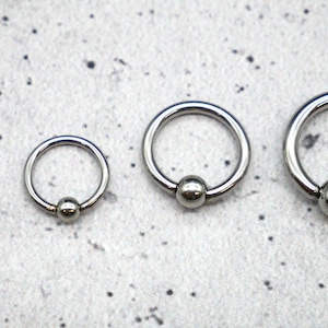 Op de afbeelding: Vier zilveren ronde ringen met een bal aan het einde van elke ring. De ringen zijn van verschillende grootte, met de kleinste ring links en de grootste ring rechts.