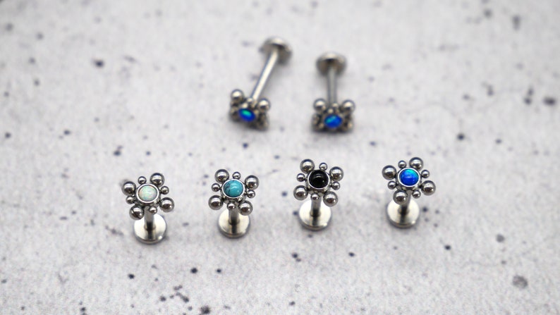 May include: Six silver body jewelry pieces with a small round gem in the center of a flower-shaped design. The gems are turquoise, black, white, and blue.