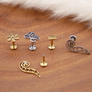 Pode incluir: Uma coleção de seis peças de joalharia corporal em metal, incluindo um pino de flor dourada, uma forma de cogumelo prateada, uma coroa dourada, um pino em forma de asa prateada, um pino em forma de asa dourada e um poste dourado plano.