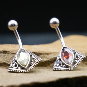 May include: Two silver belly button rings with ornate designs. One ring has a clear green gem, and the other has a red gem.