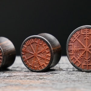 Könnte beinhalten: Drei Holzohrstecker mit einem geschnitzten Vegvisir-Symbol in rotbrauner Farbe. Die Stecker sind außen schwarz und haben eine Holzmaserung.