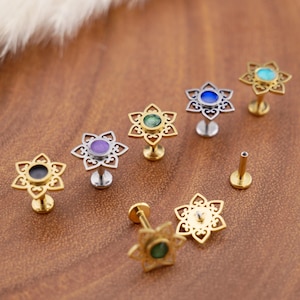 Op de afbeelding: Een collectie metalen labret-piercings met een zesbladige bloemvormige filigraan top en een centrale ronde edelsteen. De piercings zijn verkrijgbaar in goud- en zilverkleurige tinten met diverse gekleurde stenen, waaronder zwart, paars, groen, blauw en turkoois.