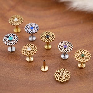 Könnte beinhalten: Eine Sammlung von Labret-Steckern mit flacher Rückseite und aufwendigen, durchbrochenen, mandalaartigen Blumenaufsätzen. Die Stecker sind in Gold- und Silbertönen gehalten und jeweils mit einem kleinen, runden, farbigen Stein in Weiß, Blau, Schwarz, Lila und Grün besetzt.