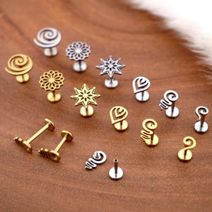 Puede incluir: Una colección de pendientes de botón variados en tonos dorados y plateados. Los diseños incluyen espirales, flores, estrellas, corazones y formas abstractas. Los pendientes se exhiben sobre una superficie marrón.