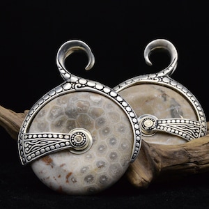 Peut inclure: Une paire de boucles d'oreilles en argent et or avec un motif de pierre fossilisée. Les boucles d'oreilles sont en forme de cercle avec un petit crochet sur le dessus. La pierre est de couleur brun clair avec un motif blanc et noir.