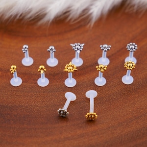 May include: A set of 12 clear plastic body jewellery pieces with different gold and silver coloured tops. The tops are in various shapes, including stars, circles, and spikes.
