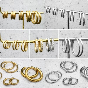 Pode incluir: Um conjunto de brincos de argola dourados e prateados. Os brincos são exibidos em um suporte. Os brincos dourados são lisos, enquanto os brincos prateados são adornados com pequenas pedras de zircônia cúbica.