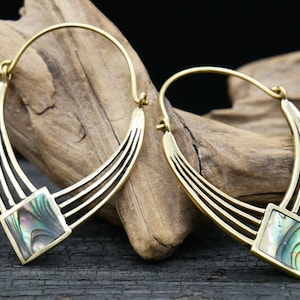 May include: A pair of gold hoop earrings with a square abalone shell inlay. The earrings have a curved design and are hanging from a wooden surface.