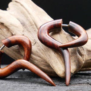 Puede incluir: Un par de dilatadores de oreja de madera con un diseño curvo único. Los dilatadores están hechos de madera marrón oscura con un tono rojizo y tienen un poste de metal plateado.