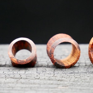 May include: Four wooden ear gauges of different sizes, with a reddish-brown color and a natural wood grain pattern. The gauges are arranged in a row on a dark, textured surface.