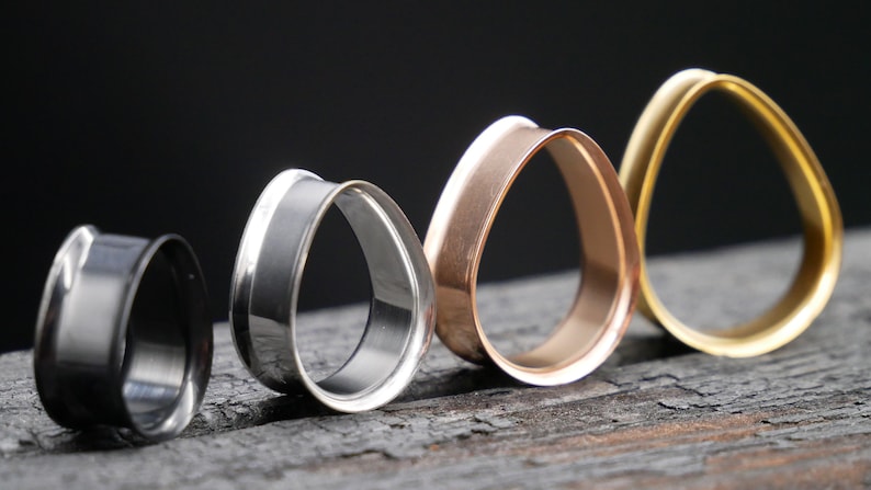 May include: Four different colored teardrop-shaped ear gauges. The gauges are made of metal and are arranged in a row from left to right. The colors are black, silver, rose gold, and gold.